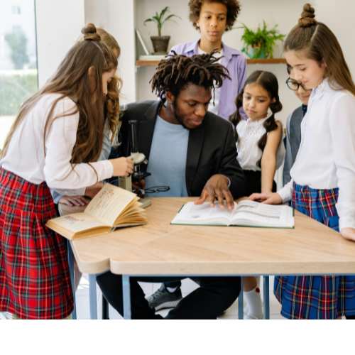 Educator teaching a group of elementary students using a microscope and reading from a lesson pack in a classroom setting.