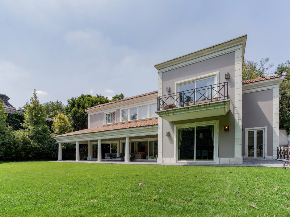 Casa en Lomas de Chapultepec, Parque Vía Reforma. Casa residencial de estilo contemporáneo con jardín amplio, fachada en tonos claros, grandes ventanales de piso a techo, terraza cubierta y balcón en planta alta, rodeada de vegetación y árboles.