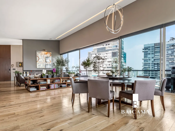 Club Residencial Bosques: Comedor moderno en un departamento de lujo con piso de madera, mesa redonda y sillas gris claro, grandes ventanales con vista a edificios contemporáneos y librero bajo con plantas decorativas.