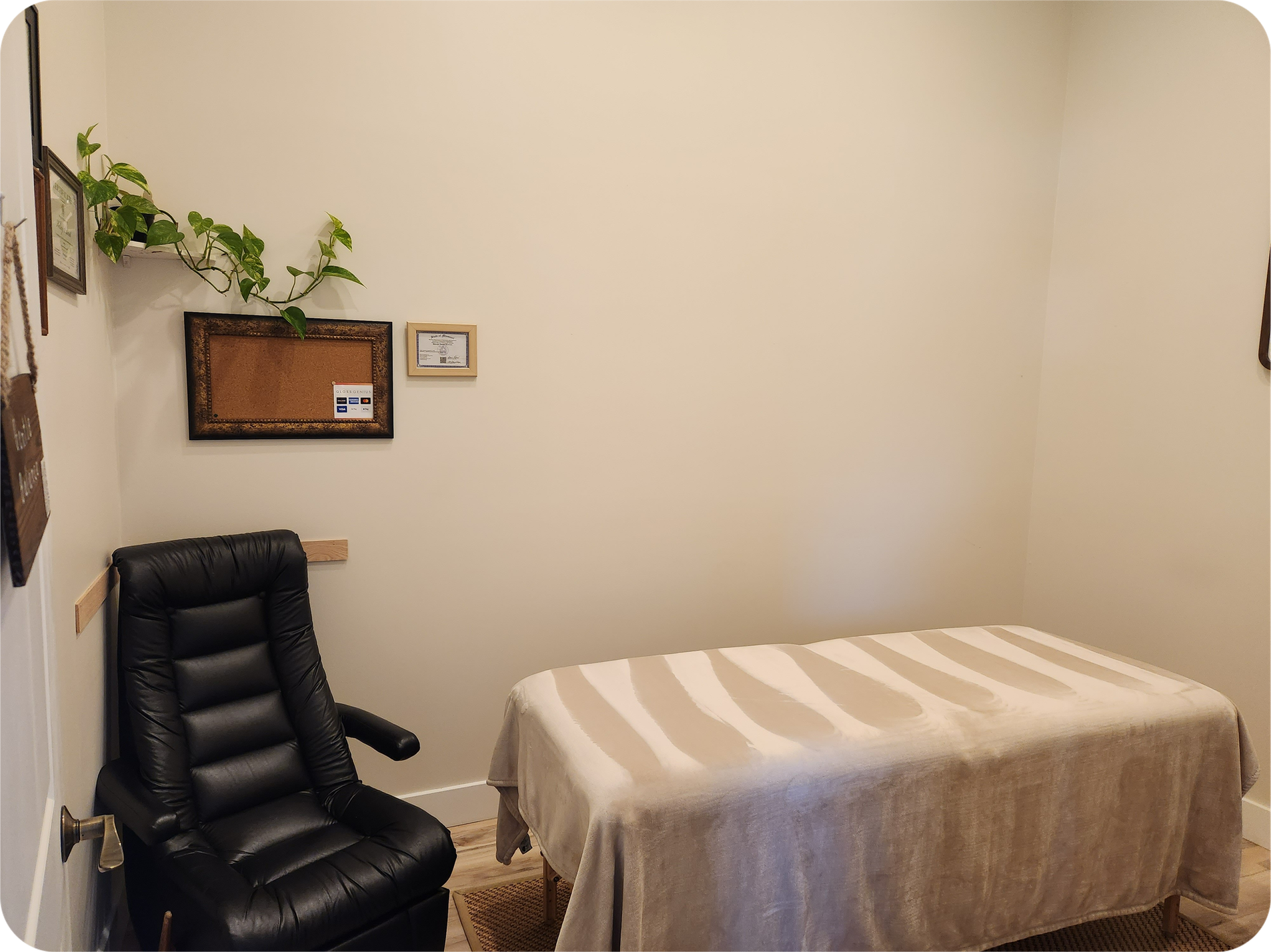 The inside of the massage room has a slim black armchair on the left and massage table with soft blanket in the center. To the right is Japanese calligraphy art and a table that a manual wheelchair can park under