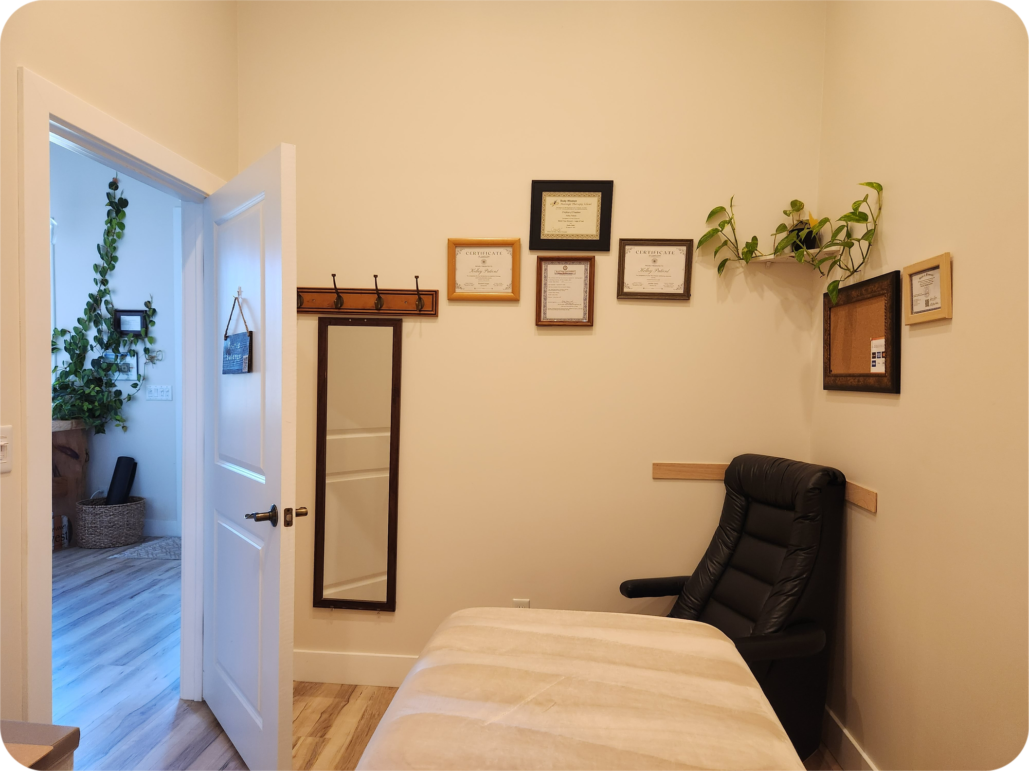 The view of the massage room from the north wall shows certifications and a happy plant on the opposite wall near the slim recliner, with massage table in the foreground