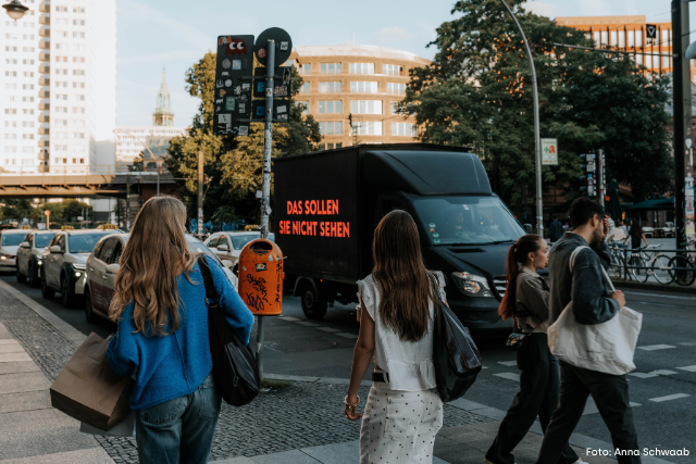 Anna und Hendrik stehen vor dem Brandenburger Tor mit dem Video-LKW und einem Schild in den Händen: