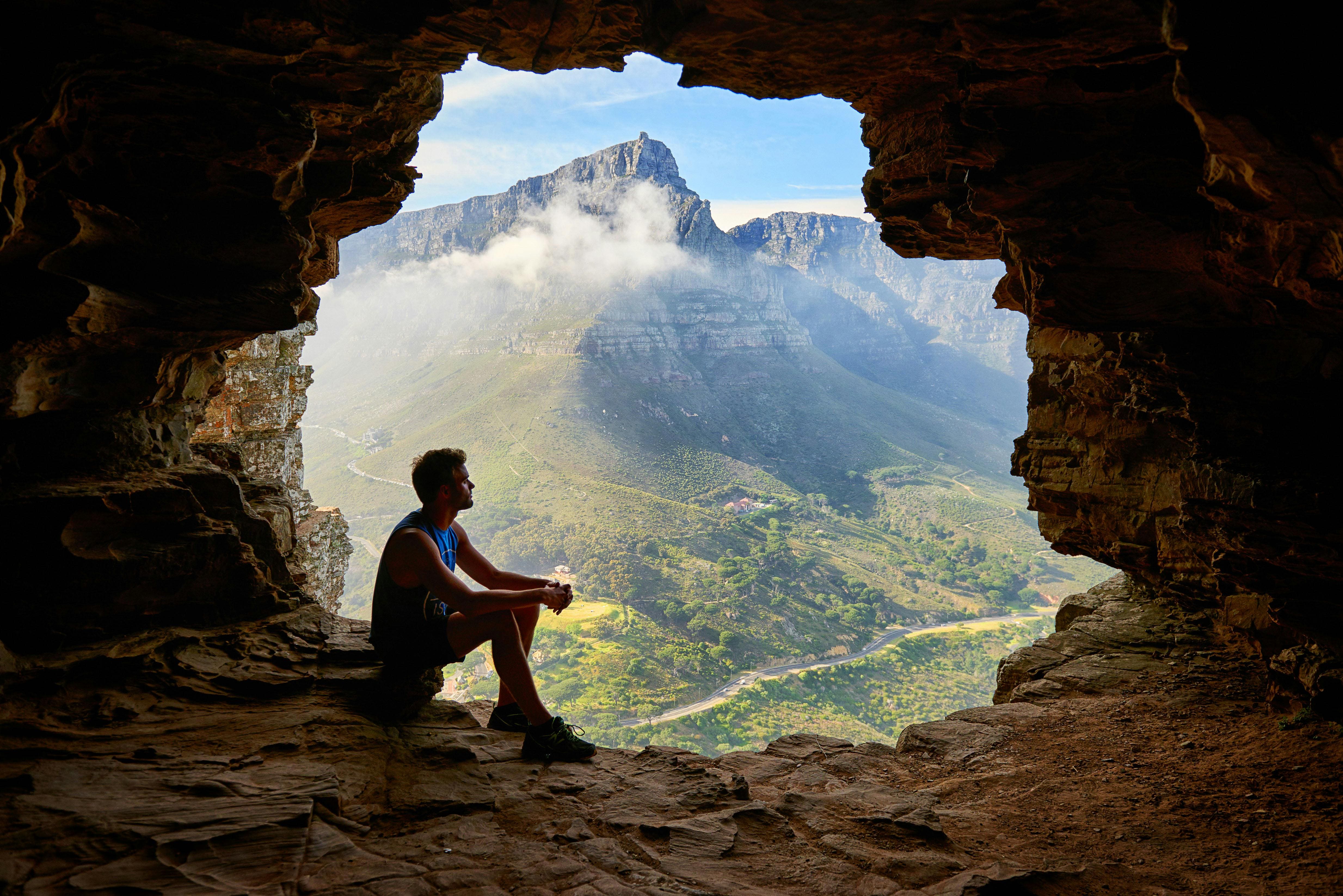 Man sitting in mountain cave opening, overlooking peaceful valley during intentional travel healing retreat