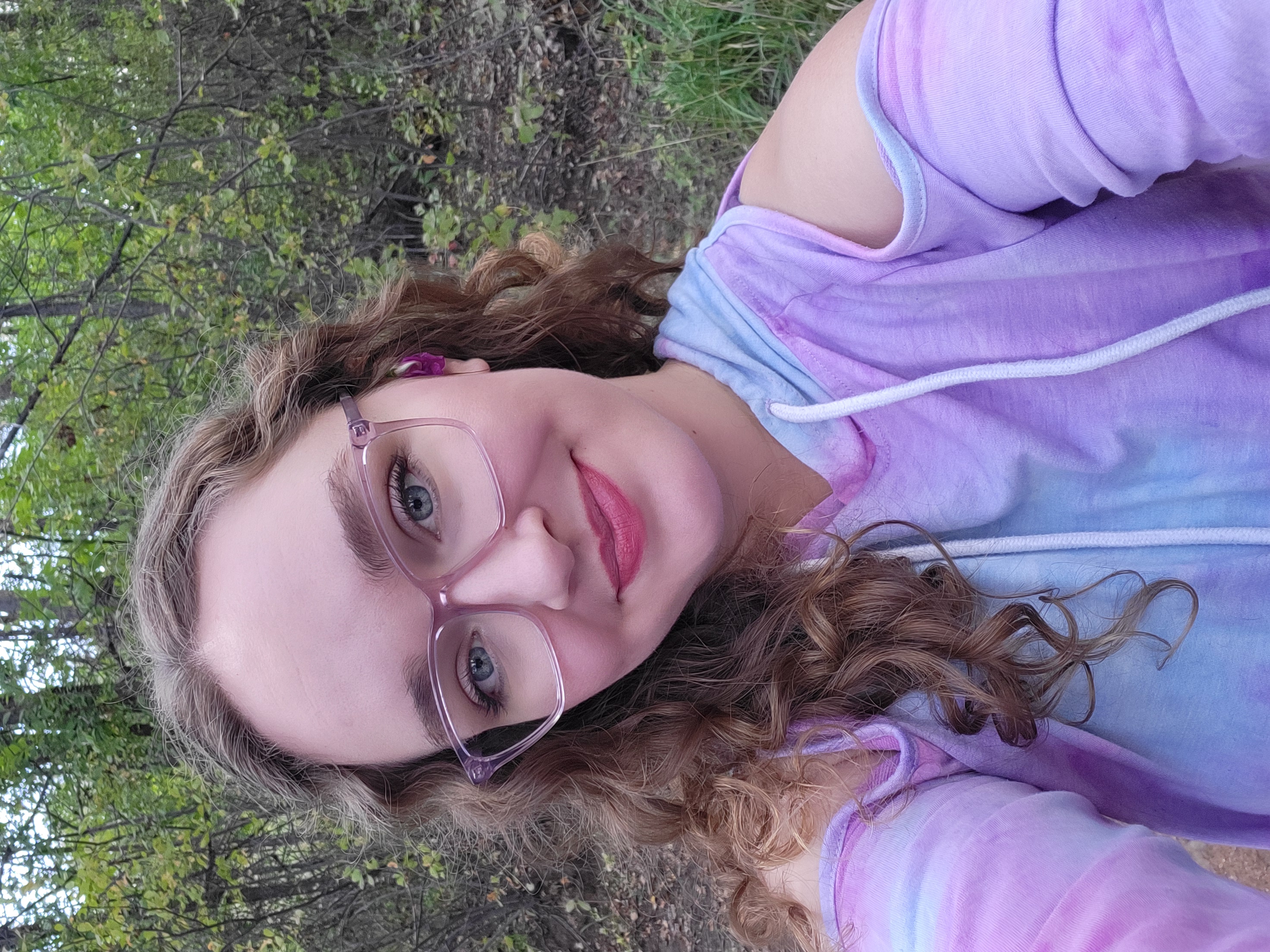 An image of a young woman with light brown, curly hair, glasses, and pink lipstick. She is looking at the camera with a peaceful smile.