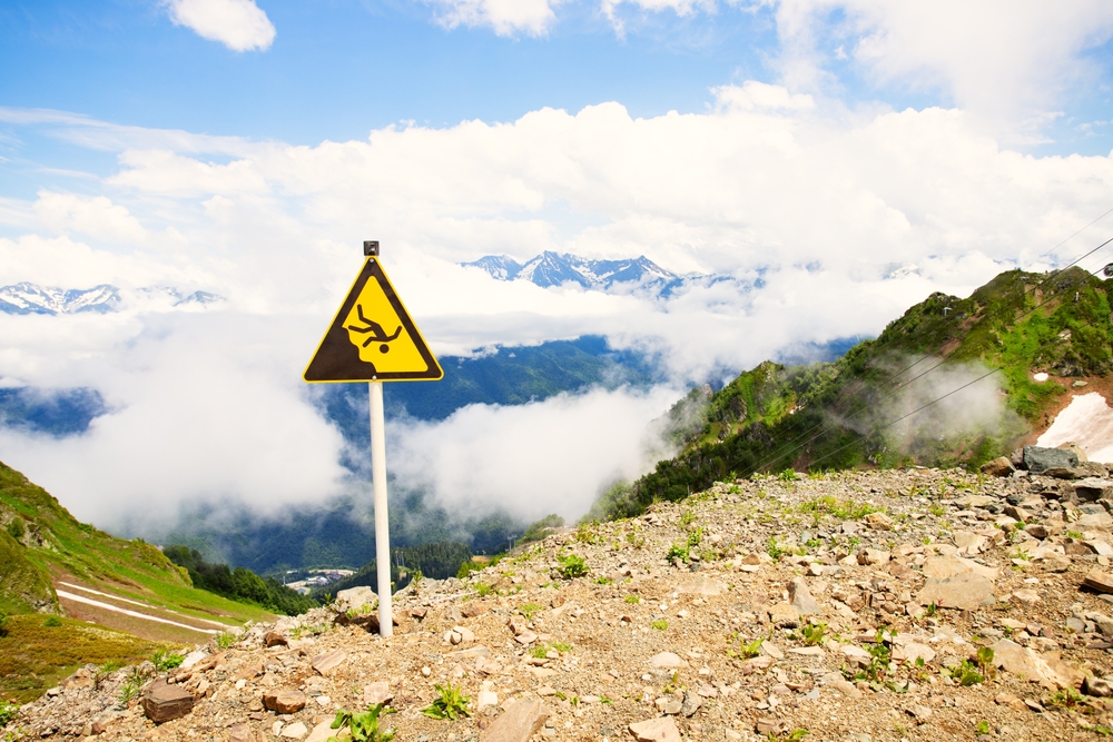 A yellow falling-hazard warning sign stands on a rocky mountain slope above clouds, with distant snowcapped peaks under a blue sky.