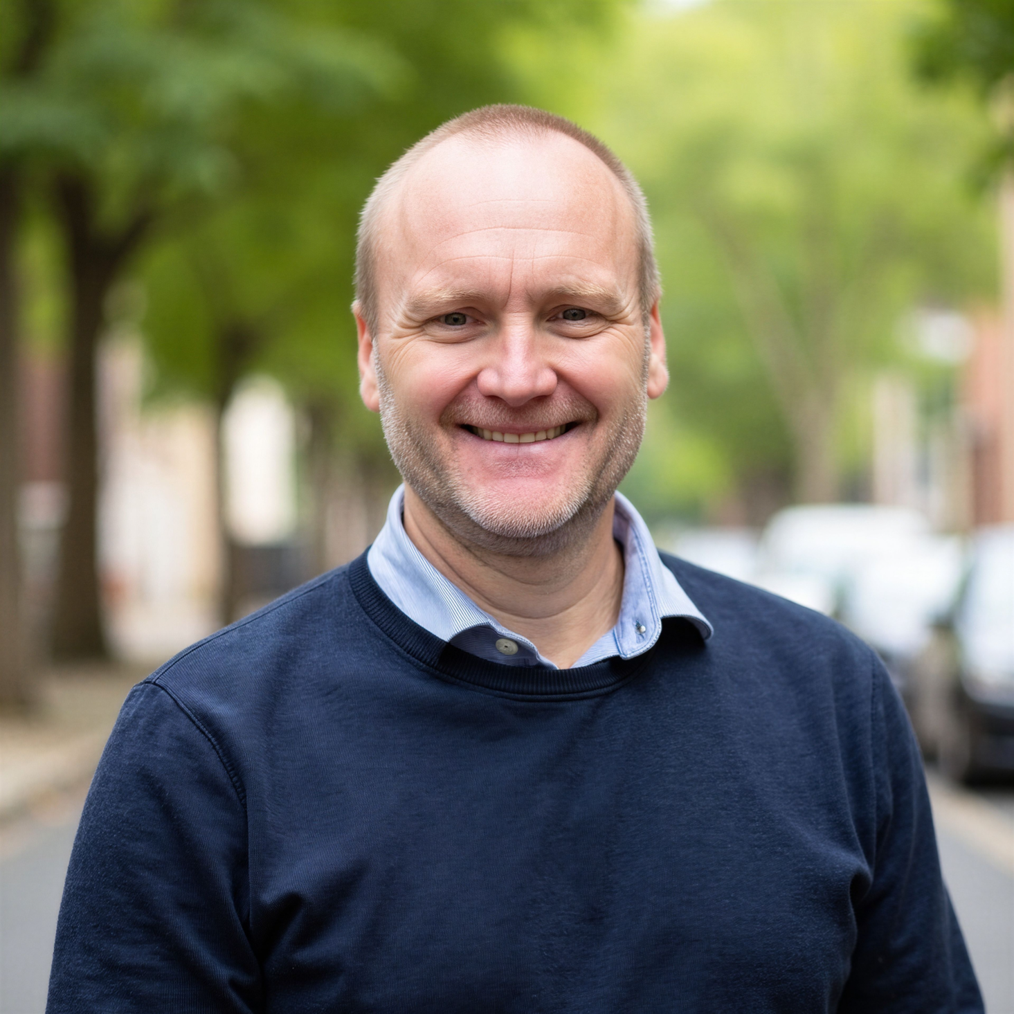 Chad Brown smiling in a navy blue sweater over a light blue collared shirt, standing outdoors on a tree-lined street with a softly blurred background.