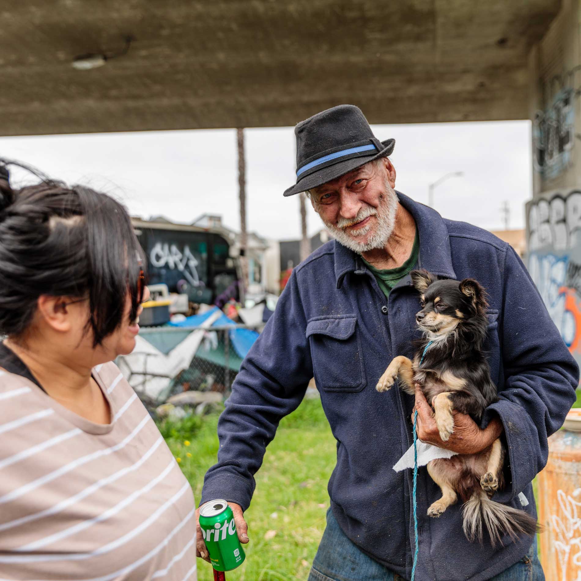 An old man with a white beard and moustache, grey fedora hat with cornflower blue band holds a black and tan longhaired chihuahua and a Sprite can. A woman laughs looking at him in the foreground, wearing a tan and white striped shirt.