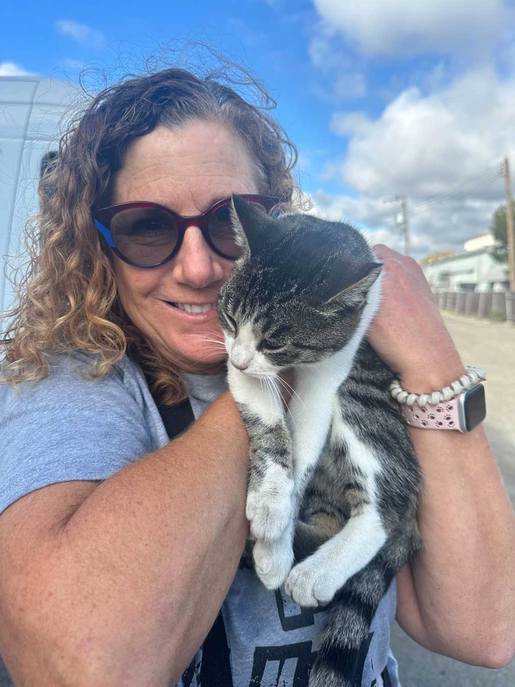 A HAPI volunteer holds up a cat, smiling at the camera. The volunteer is female, white, wearing a heathered grey t-shirt, a pink watch, and a white beaded bracelet. The cat is a grey tabby with white points.