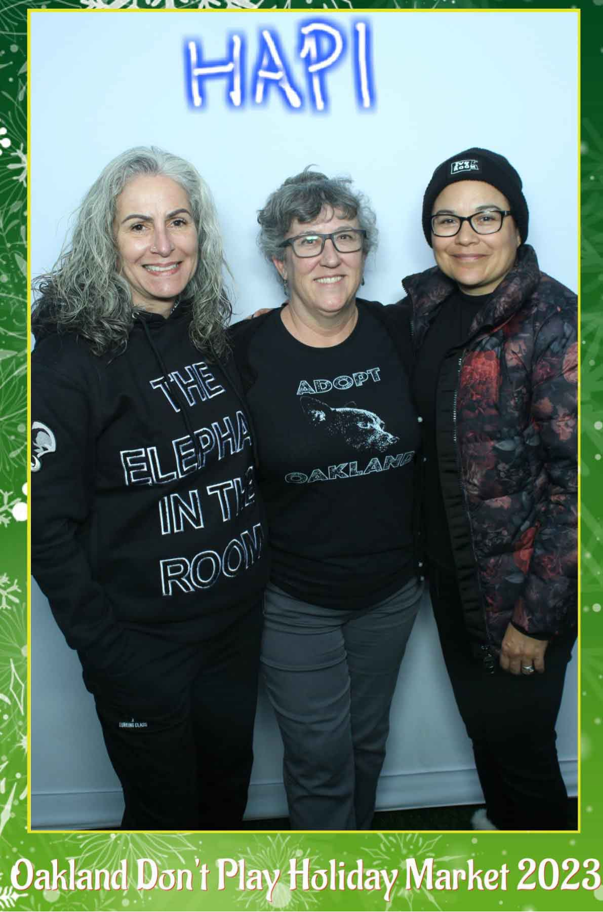 Three HAPI volunteers pose together for a photo at Oakland Don't Play, with a green holiday frame around the photo, saying 