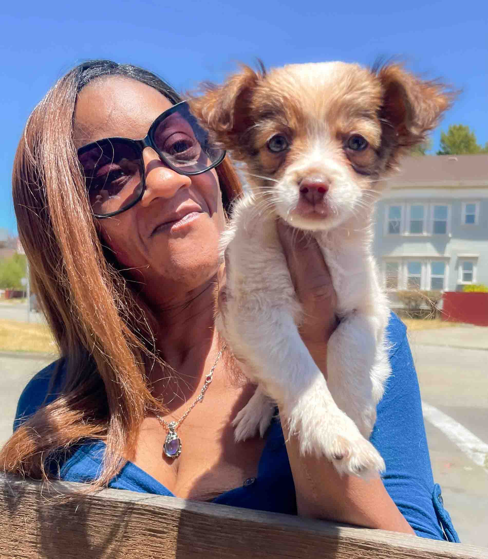 A woman, probably middle aged with caramel colored skin and long, straight red hair holds a small puppy against her shoulder. She wears a bright blue low cut shirt and necklace, and her puppy is white with tan ears and spots over its eyes.