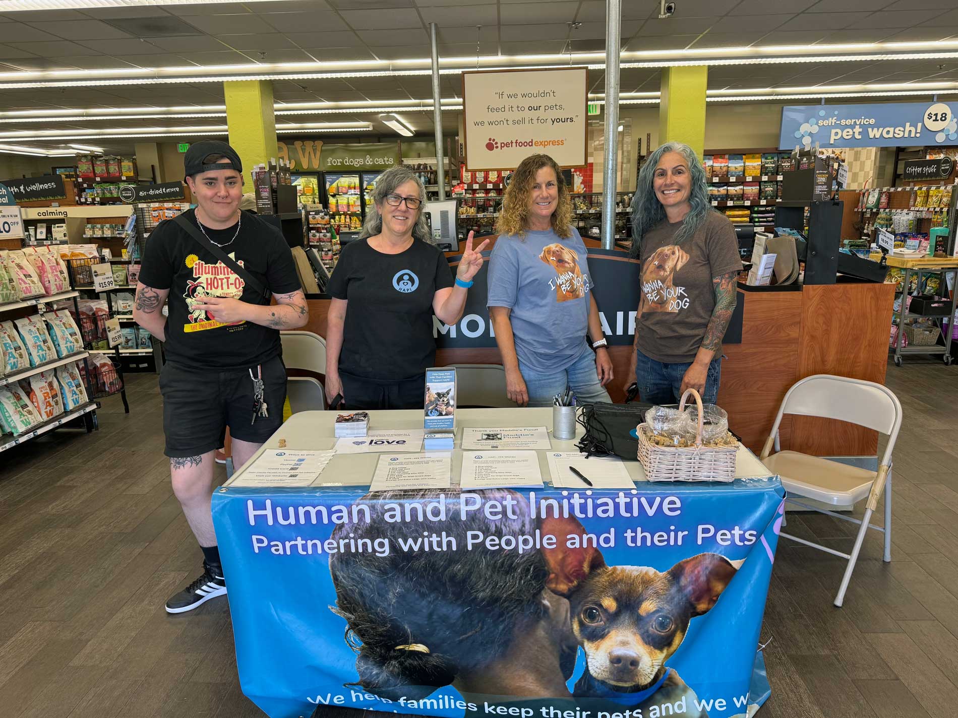 HAPI Board members Briana, Pam Sharon, and Meg stand in front of a HAPI informational table, inside of a Petfood Express store. A blue banner hanging from the table says, 