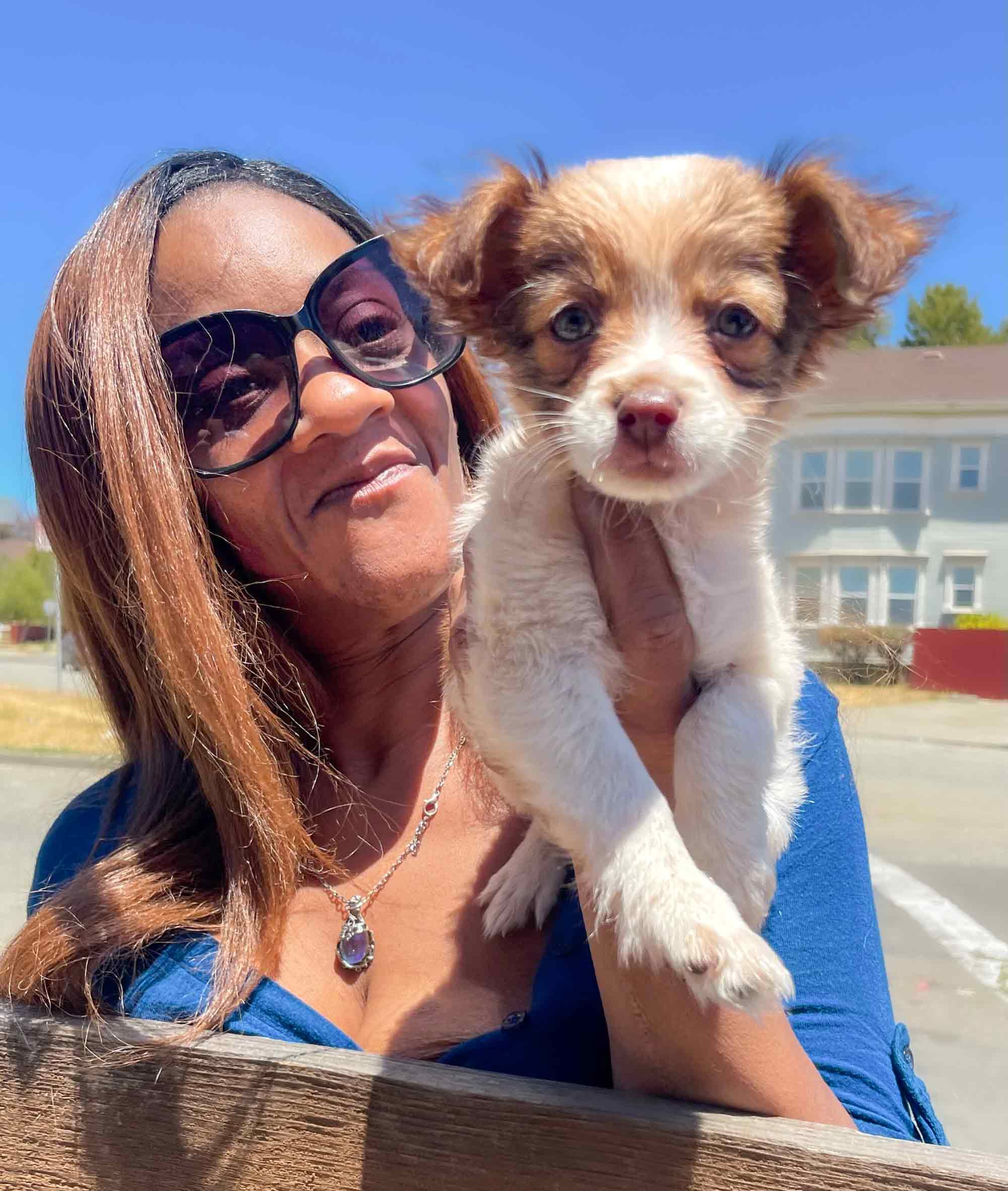 A woman, probably middle aged with caramel colored skin and long, straight red hair holds a small puppy against her shoulder. She wears a bright blue low cut shirt and necklace, and her puppy is white with tan ears and spots over its eyes.