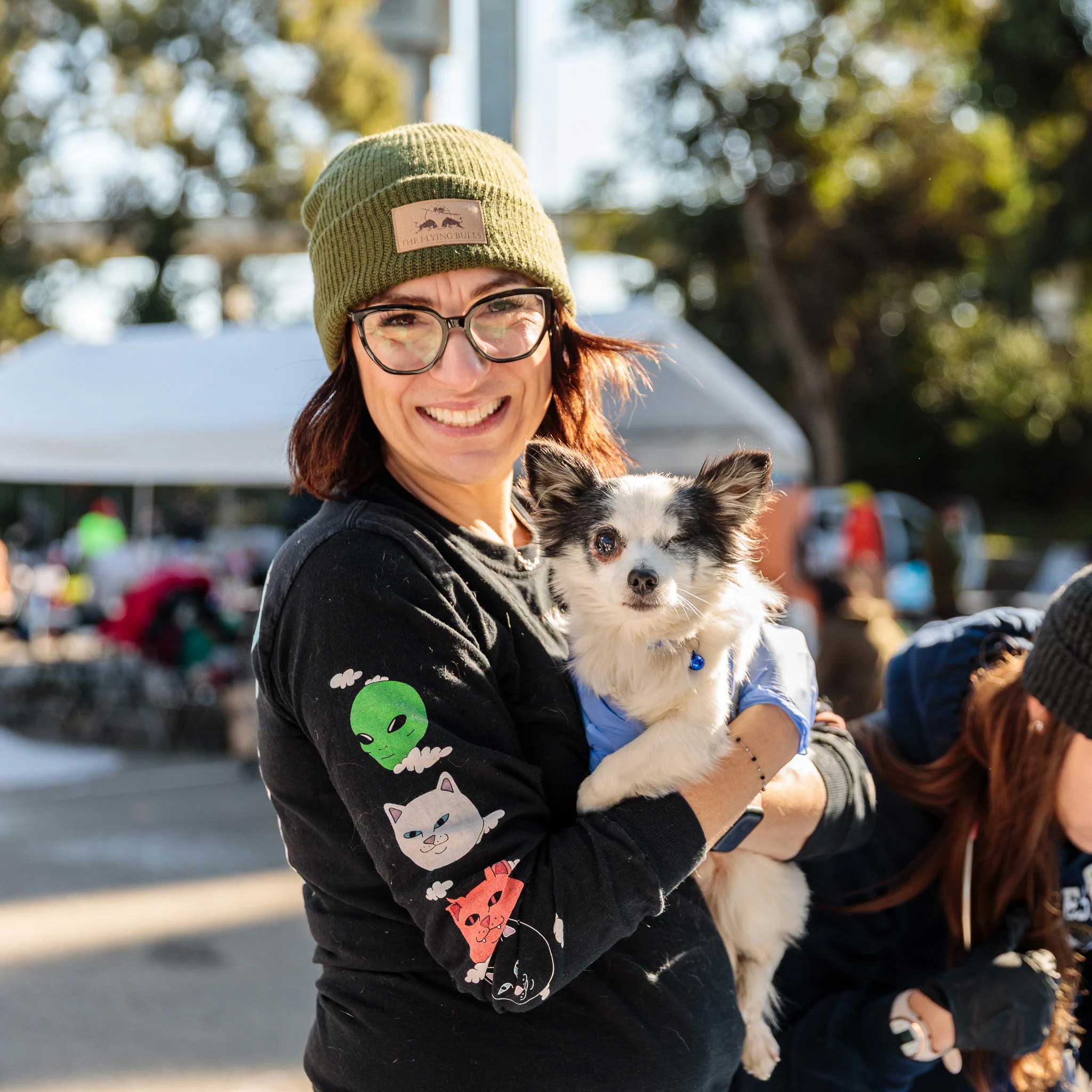 A volunteer holds a small long-haired dog, with pointy grey ears. She is a white woman wearing glasses and a dusty green beanie, a black top with little colorful cats down the long sleeve. She boasts a huge smile while the dog looks at the camera.