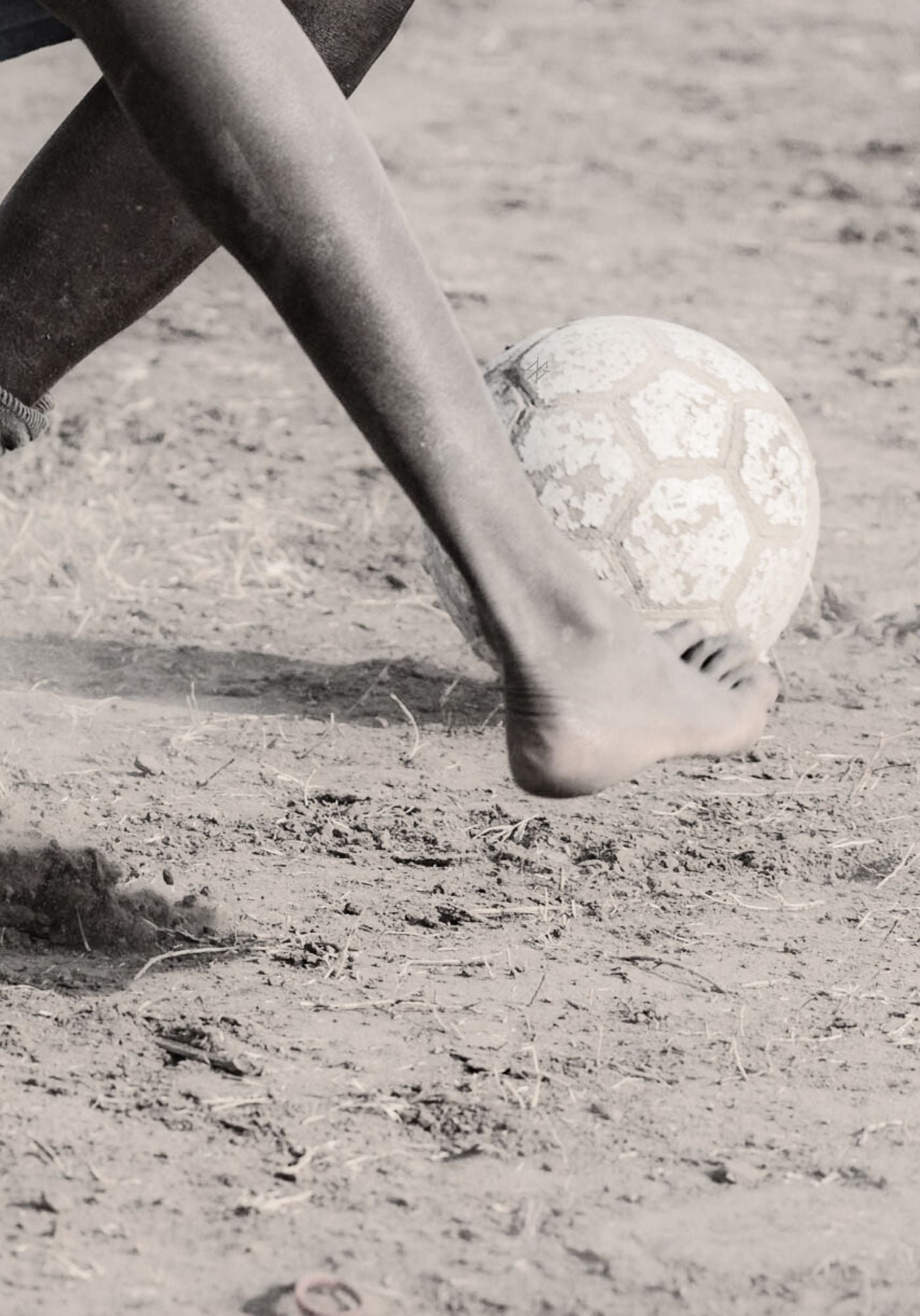 A rural football player's bare foot kicking a soccer ball on a dirt field.