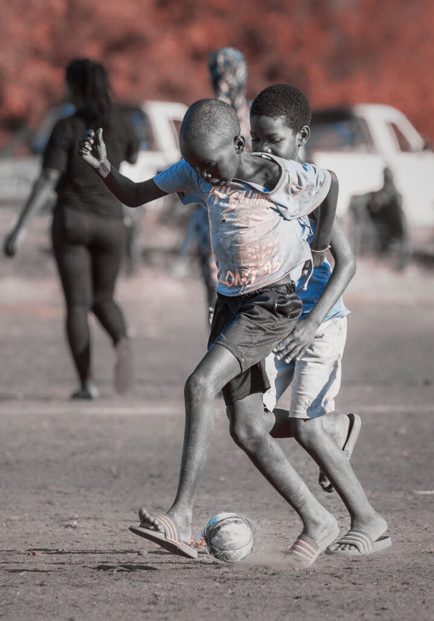 Two children playing soccer with a handmade ball, on a rural dirt field with spectators in the background.