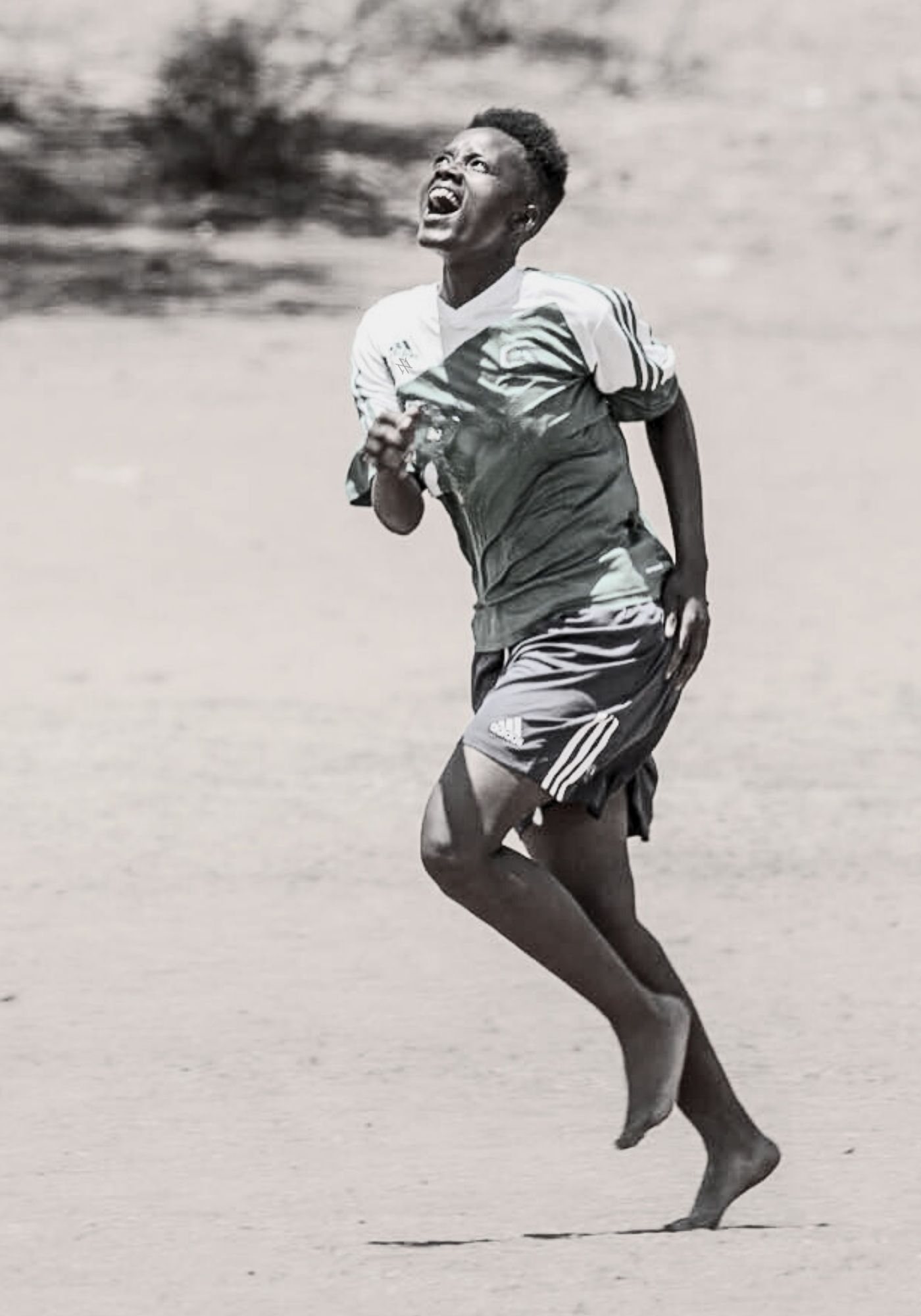 A female player in a soccer uniform running barefoot on a rural dirt pitch and shouting with their head tilted back. Celebrating a goal.