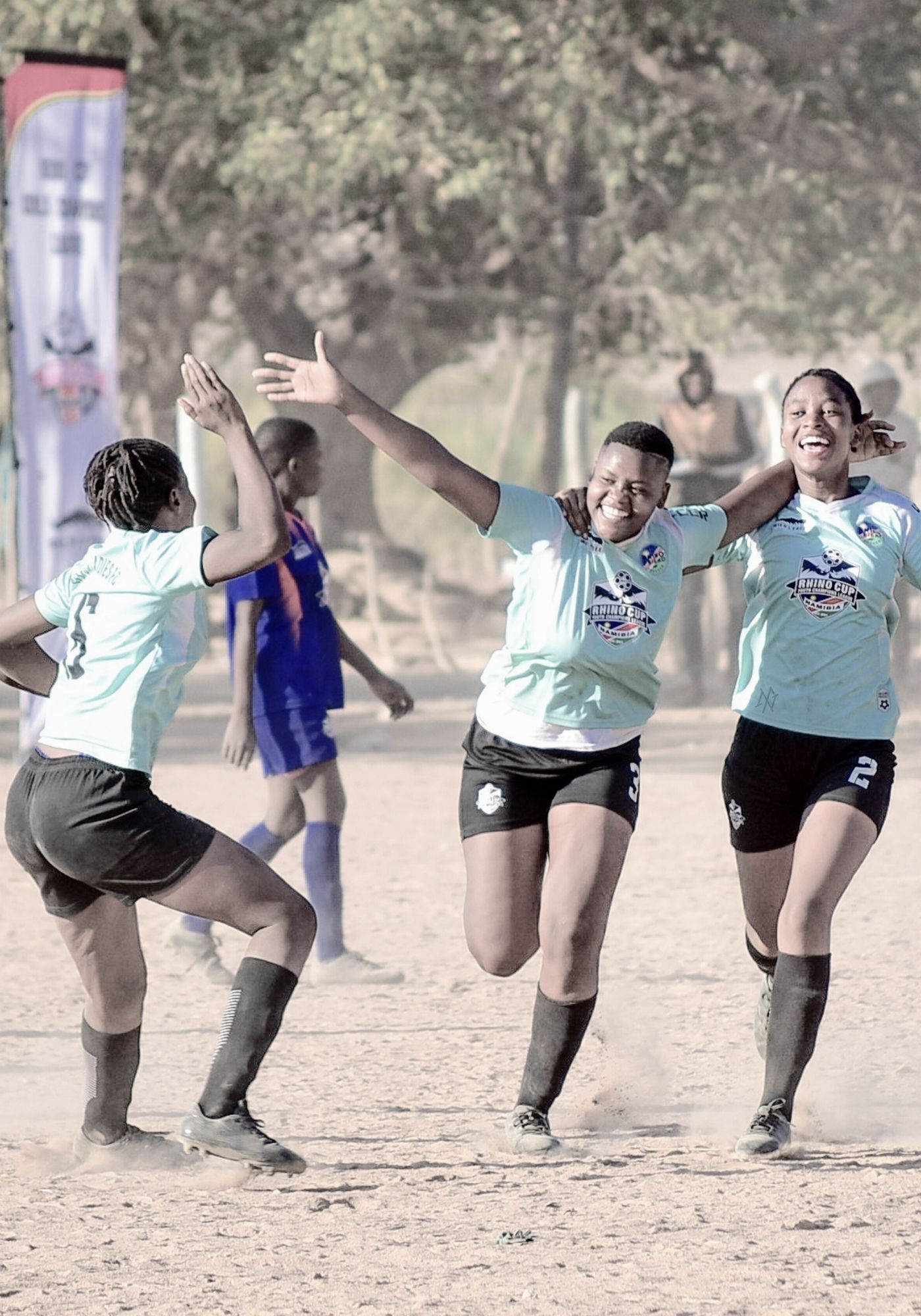 Two children playing soccer with a handmade ball, on a rural dirt field with spectators in the background.