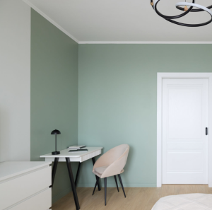 Minimalist home office corner with sage green accent wall, white desk, modern black lamp, and blush pink chair in a cozy bedroom.