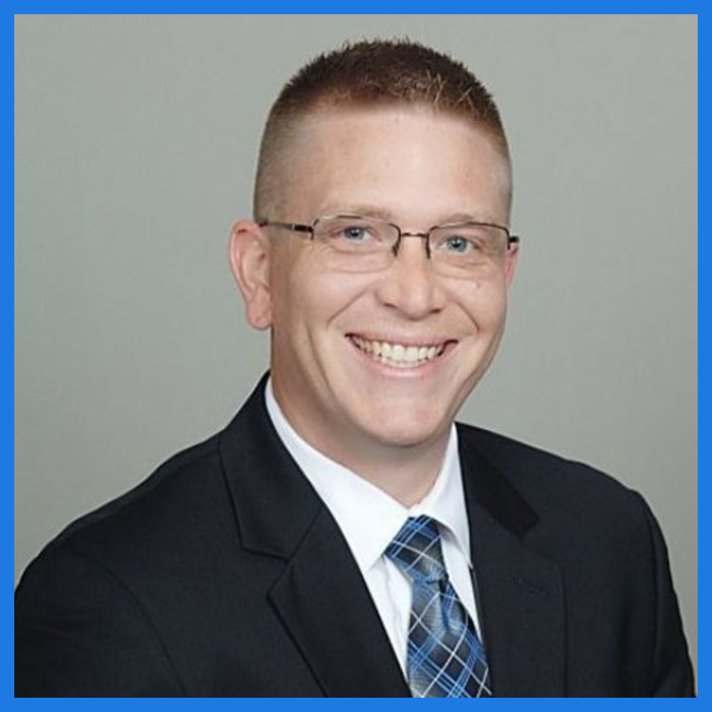 Professional headshot of a smiling man with short blond hair and horn-rimmed glasses, wearing a white dress shirt, blue suitcoat and a black and blue tie