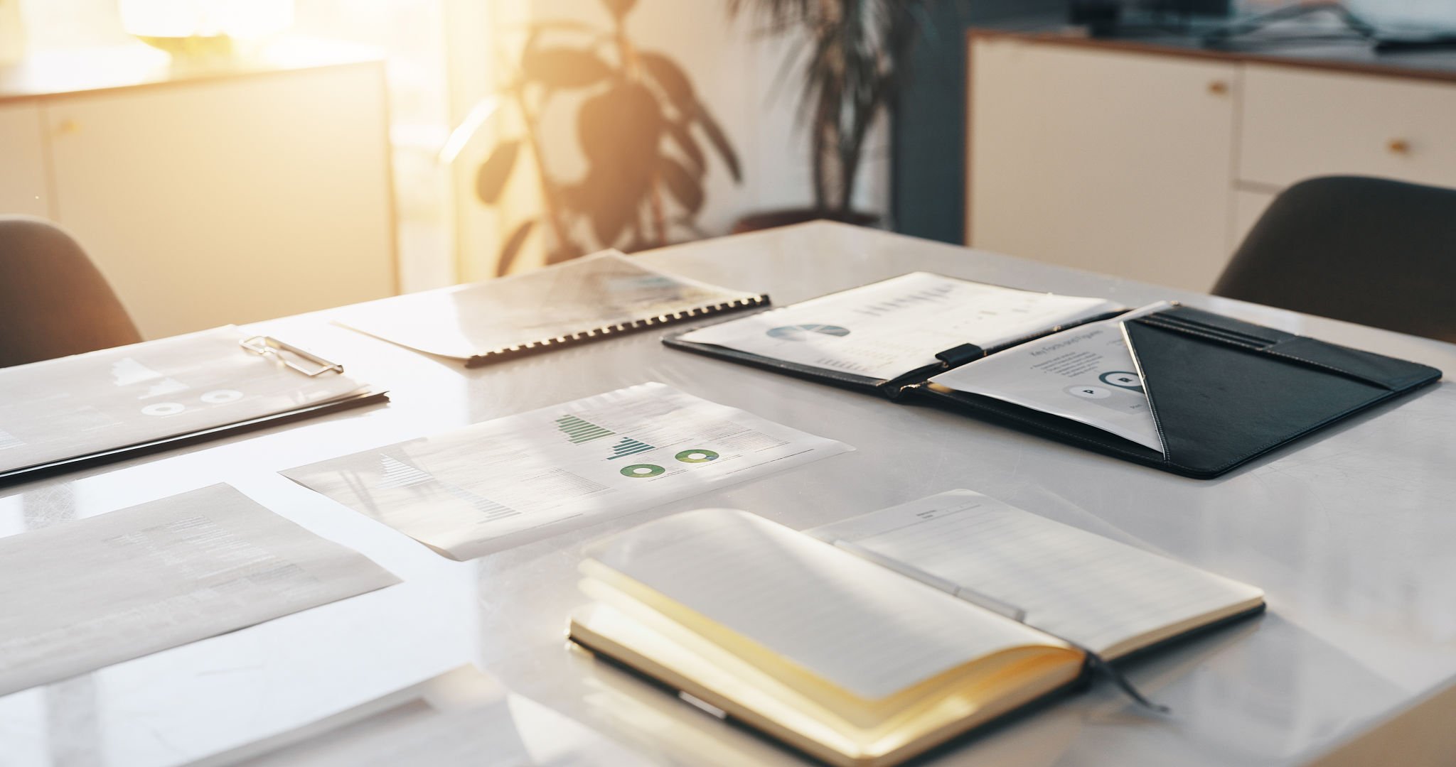 White marble table with books and portfolios neatly piled for a business meeting.  A soft warm glow of sunlight is cascading through the window and casting beautiful shadows from the green plants in the room.