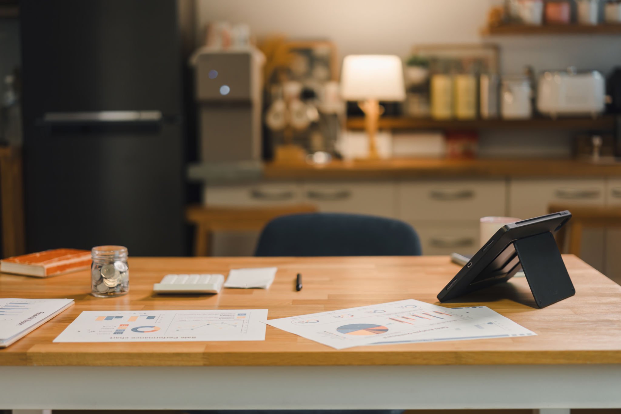 Wooden kitchen table with documents and an open tablet, promoting efficiency of work.  In the background is a slightly blurred kitchen space, with neatly organized jars and pots, creating harmony between the work space and the living space.