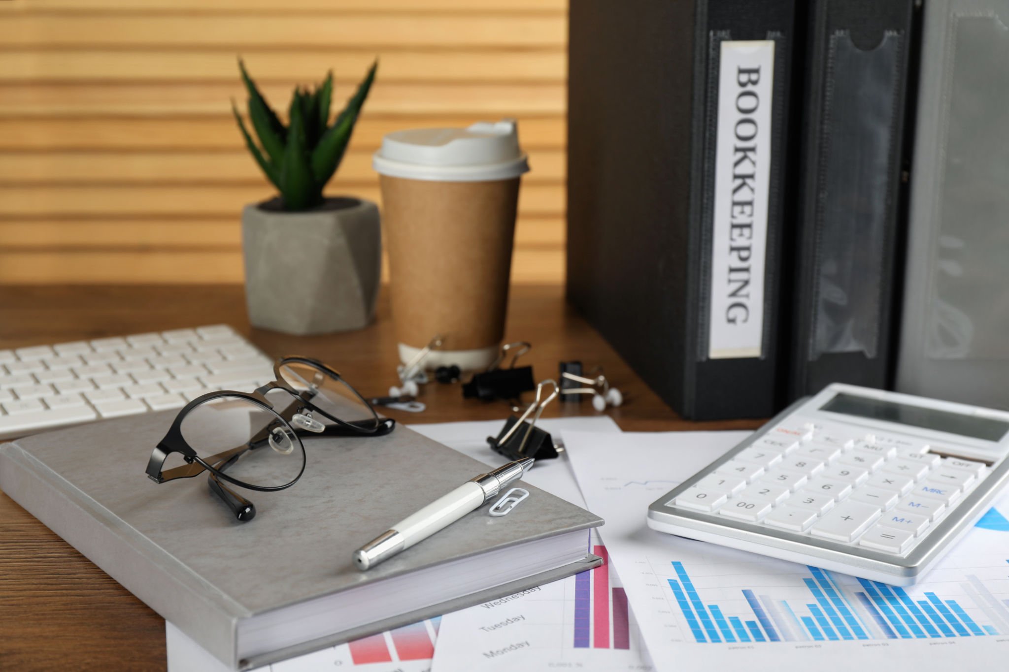 A wooden desktop with notebooks, binders, a calculator, a pen, a pair of black reading glasses, binder clips and paper clips, a cup of coffeehouse coffee and a small potted aloe plant on top of financial documents displaying graphs and figures