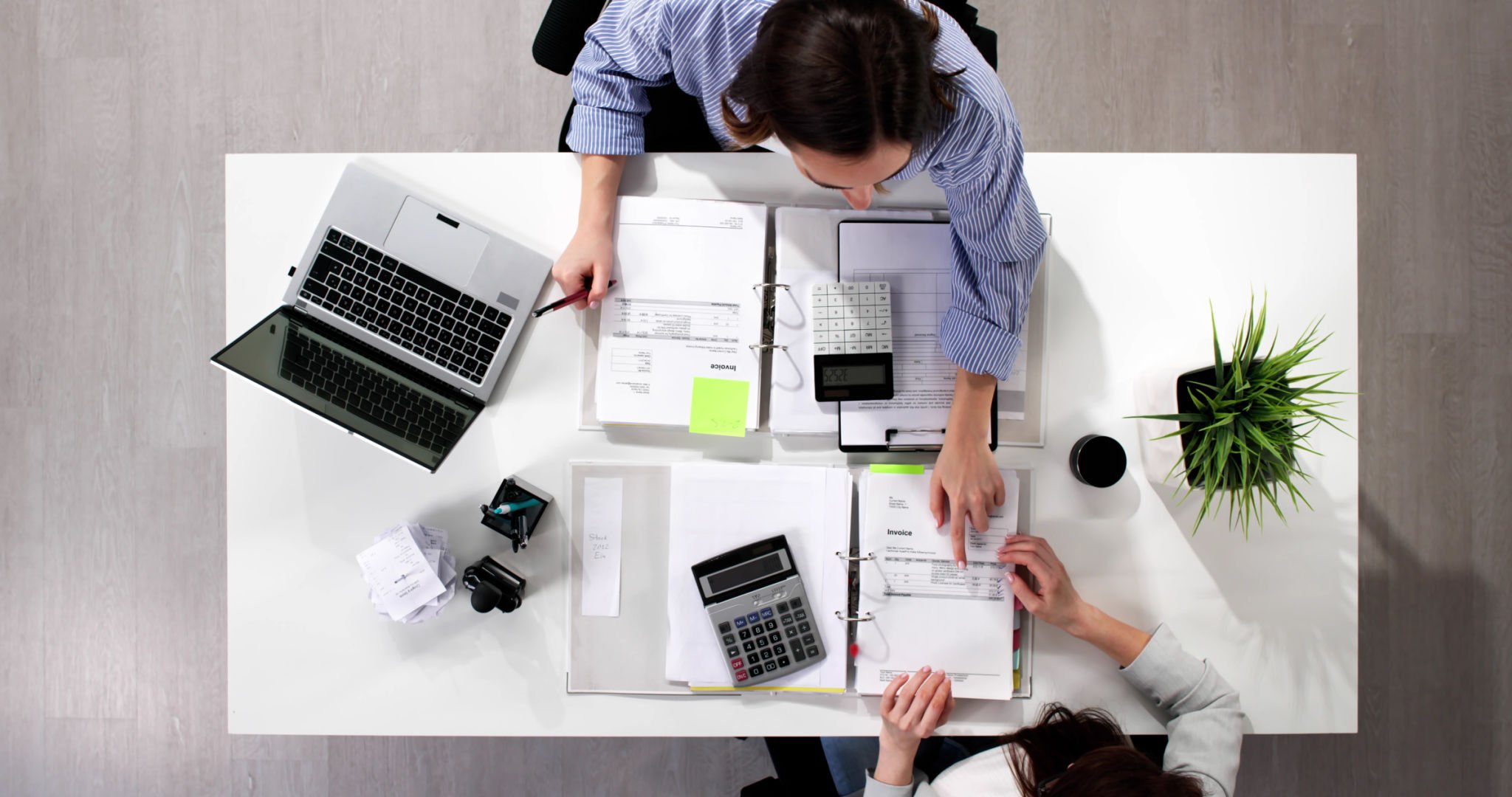 A "looking down" shot of a white table on hardwood flooring with a laptop, binders, calculators, potted plant, pens and papers.  There are two females sitting across from each other, working together on the financial reports.
