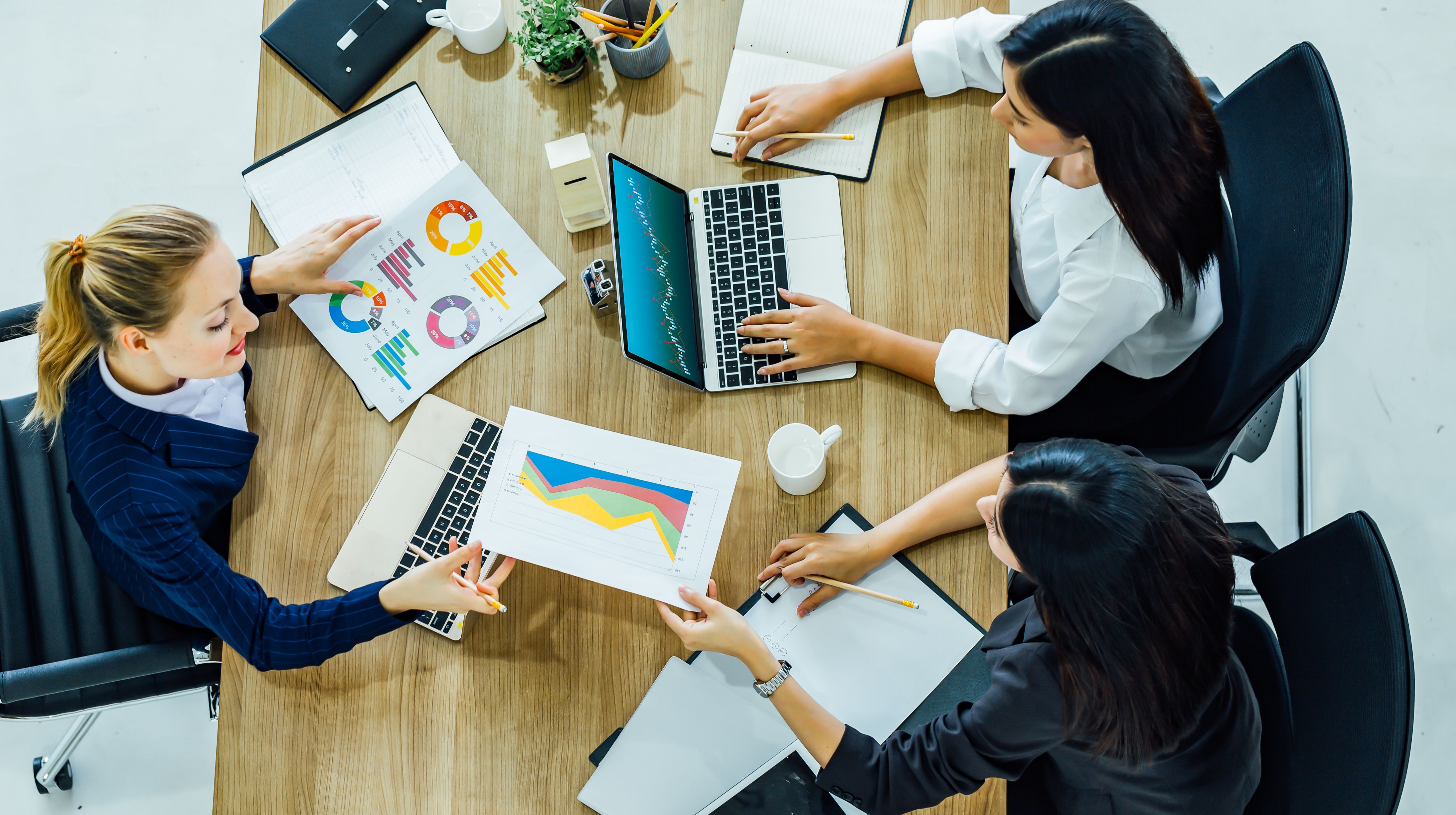 Three women sit around a table looking at printed reports and a computer screen.
