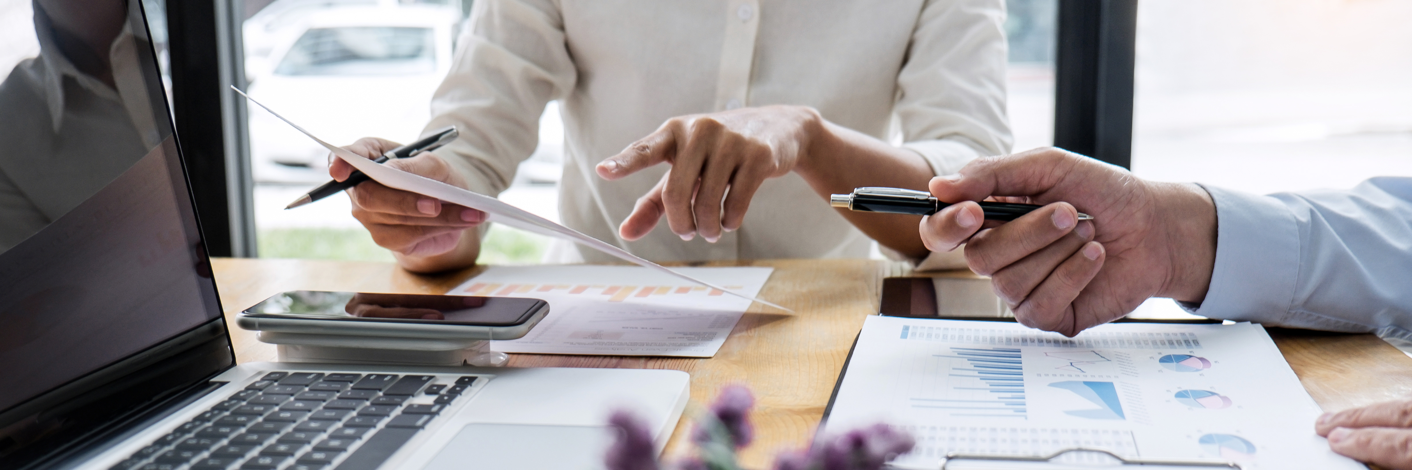 Two colleagues point at a report in one of their hands while sitting at a desk with a computer.