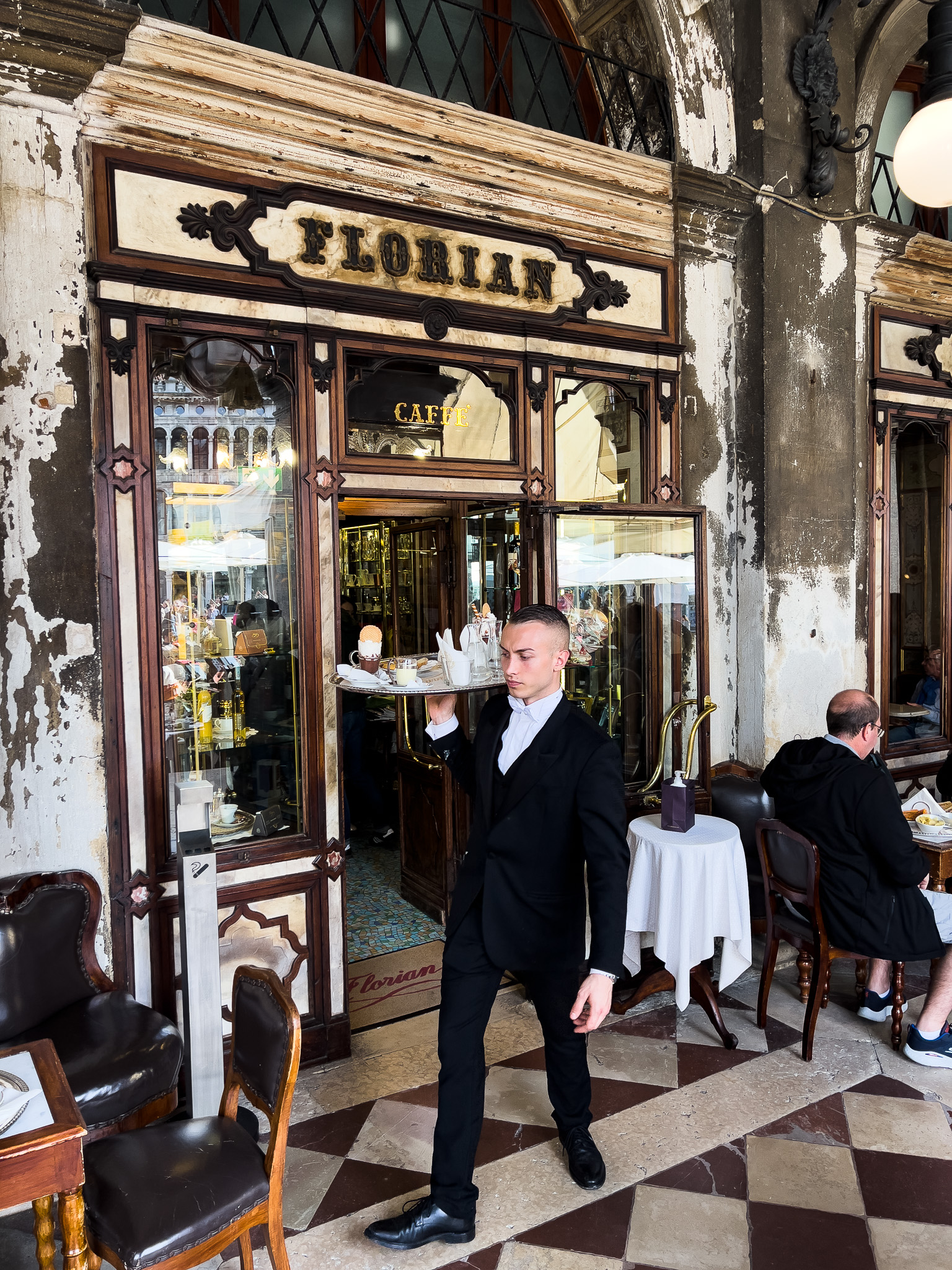 Zdjęcie pokazuje elegancko ubranego kelnera z Caffe Floriant, który niesie tacę z napojami i deserami