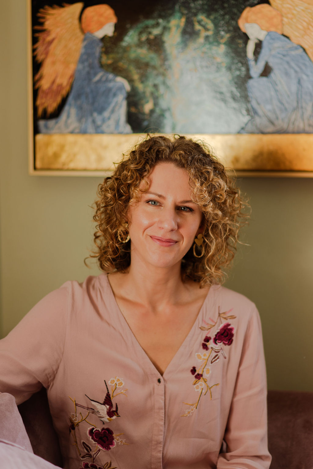 Picture of the artist smiling and with curly hair and wearing a top with birds on it and sat in front of a painting