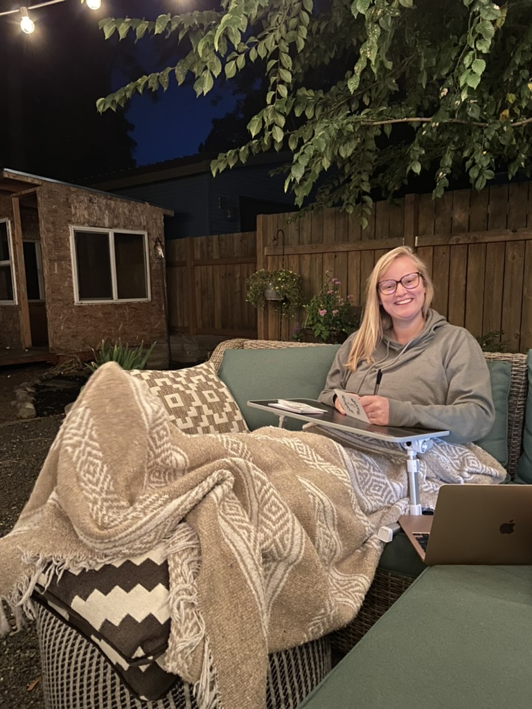 Erika is sitting on an outdoor couch smiling at the camera. She is writing on a lap desk and using a blanket. The background is a fence and some flowers.