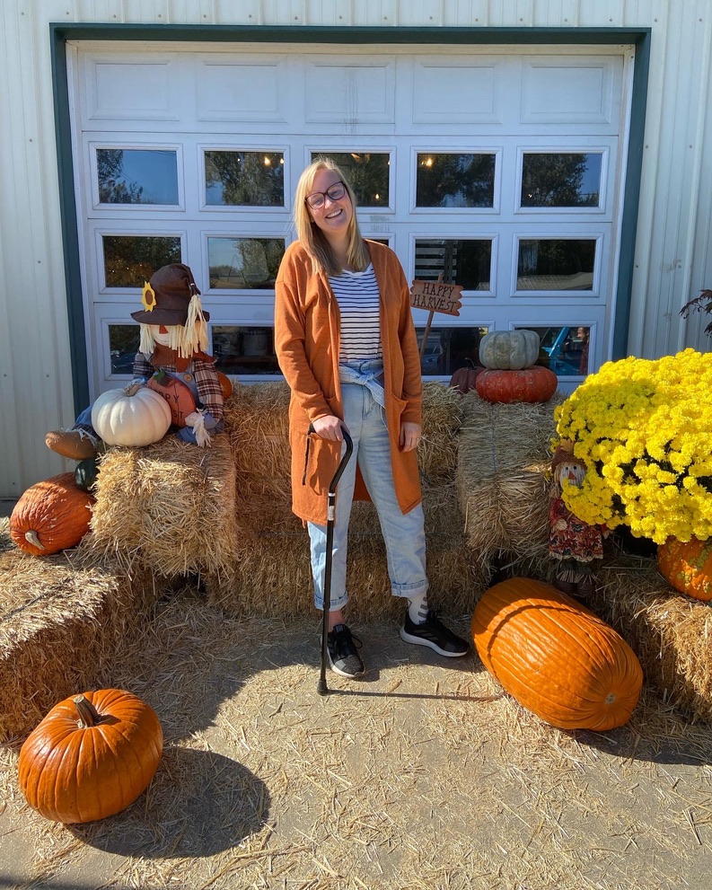 Erika is at a pumpkin patch smiling at the camera. She standing using her cane. The background is of hay, pumpkins, flowers, and a scarecrow.