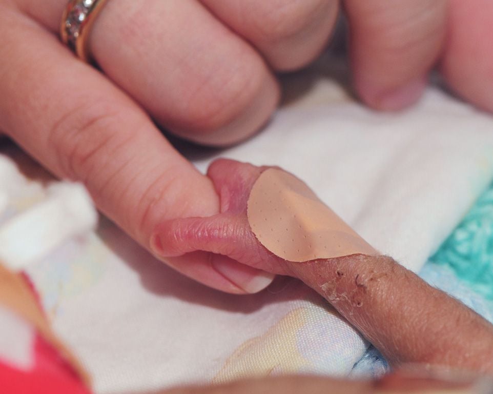 Zbliżenie na kruchą dłoń wcześniaka z małym, okrągłym opatrunkiem na palcu, delikatnie trzymaną przez dłoń dorosłej osoby – symbol opieki i bezpieczeństwa w Ośrodku Otulinka.
