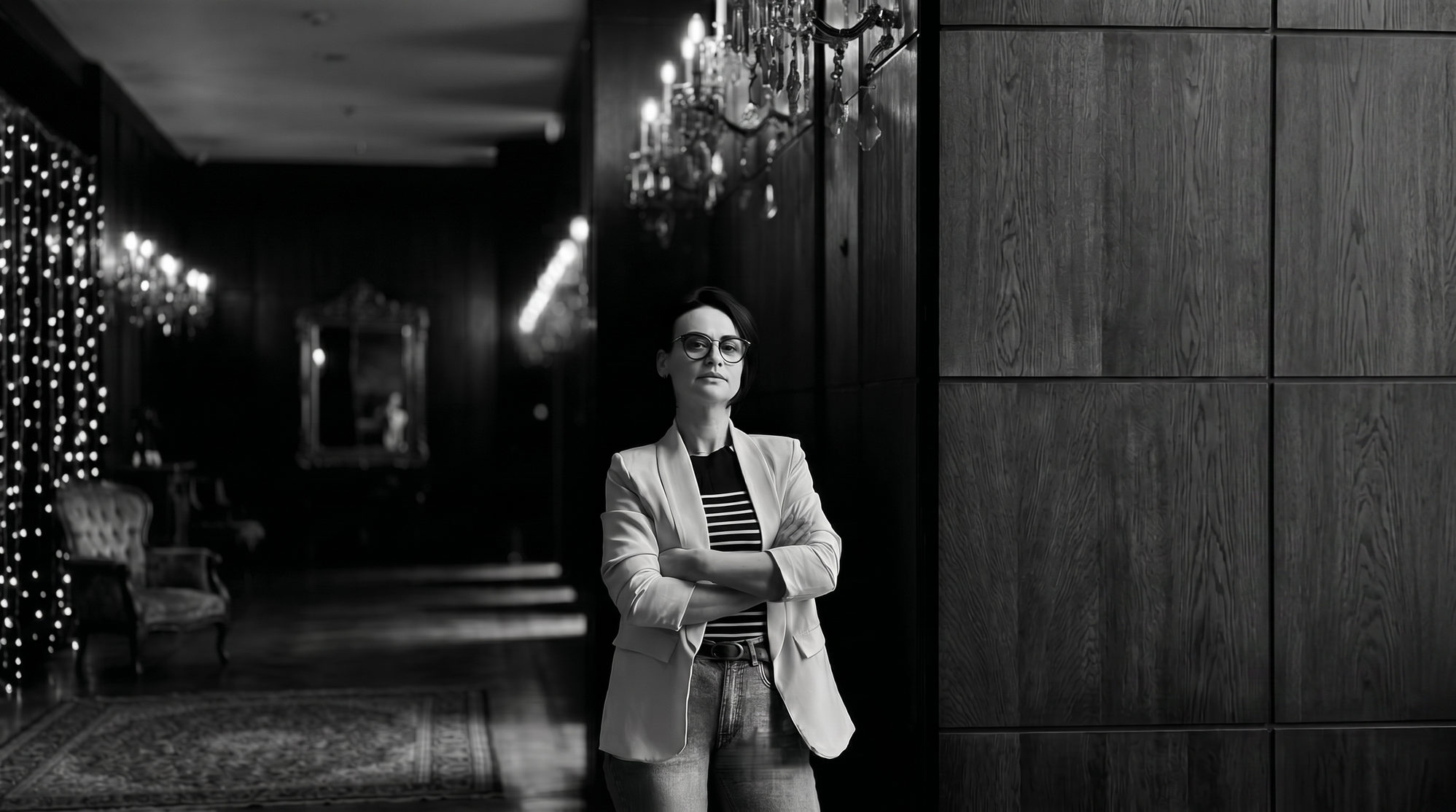 Mirela Bocanet wearing glasses and a blazer, standing in a dimly lit hallway lit by chandeliers, illustrating her journey from human strategy to AI leadership.