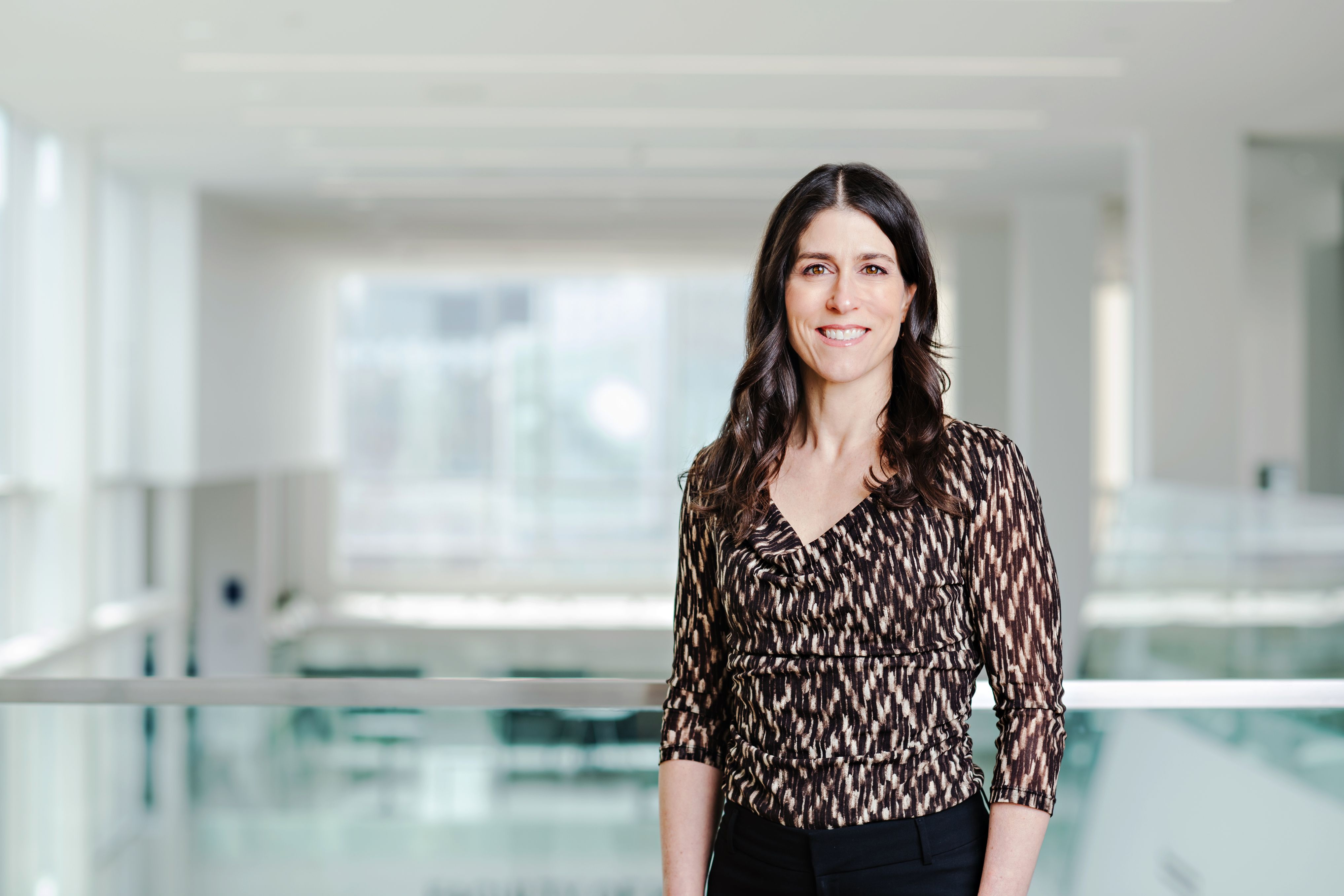 A picture of Lora Giangregorio standing against a railing on the second floor of bright academic building with white walls and glass railings. She is a white mid-forties female with long brown hair, wearing a brown and beige printed shirt and black pants.