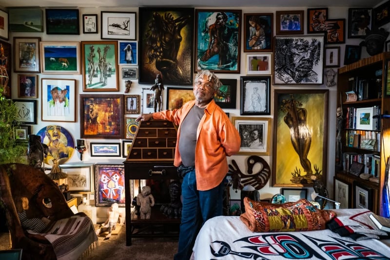 mccoy stands next to a wooden desk in a room where nearly every inch of wall is covered by art