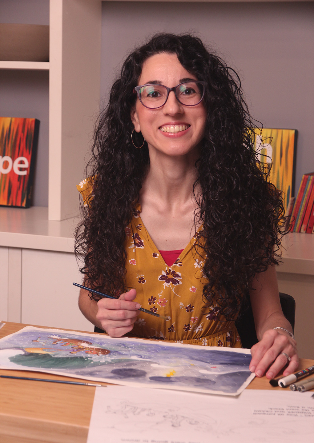 Illustrator smiling, sitting at her desk with a paintbrush in her hand and a painting of Jesus calming the storm on the desk