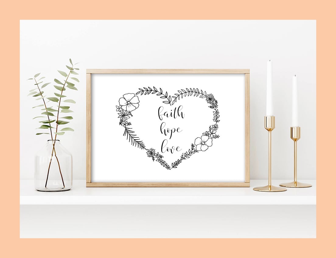 Art print with Faith Hope Love in a handwritten-style script written inside a black-and-white floral heart in a wooden frame/ The frame is propped up against the wall on a shelf, in between a glass vase with green leaves on the left and two gold candleholders with white candles on the right.