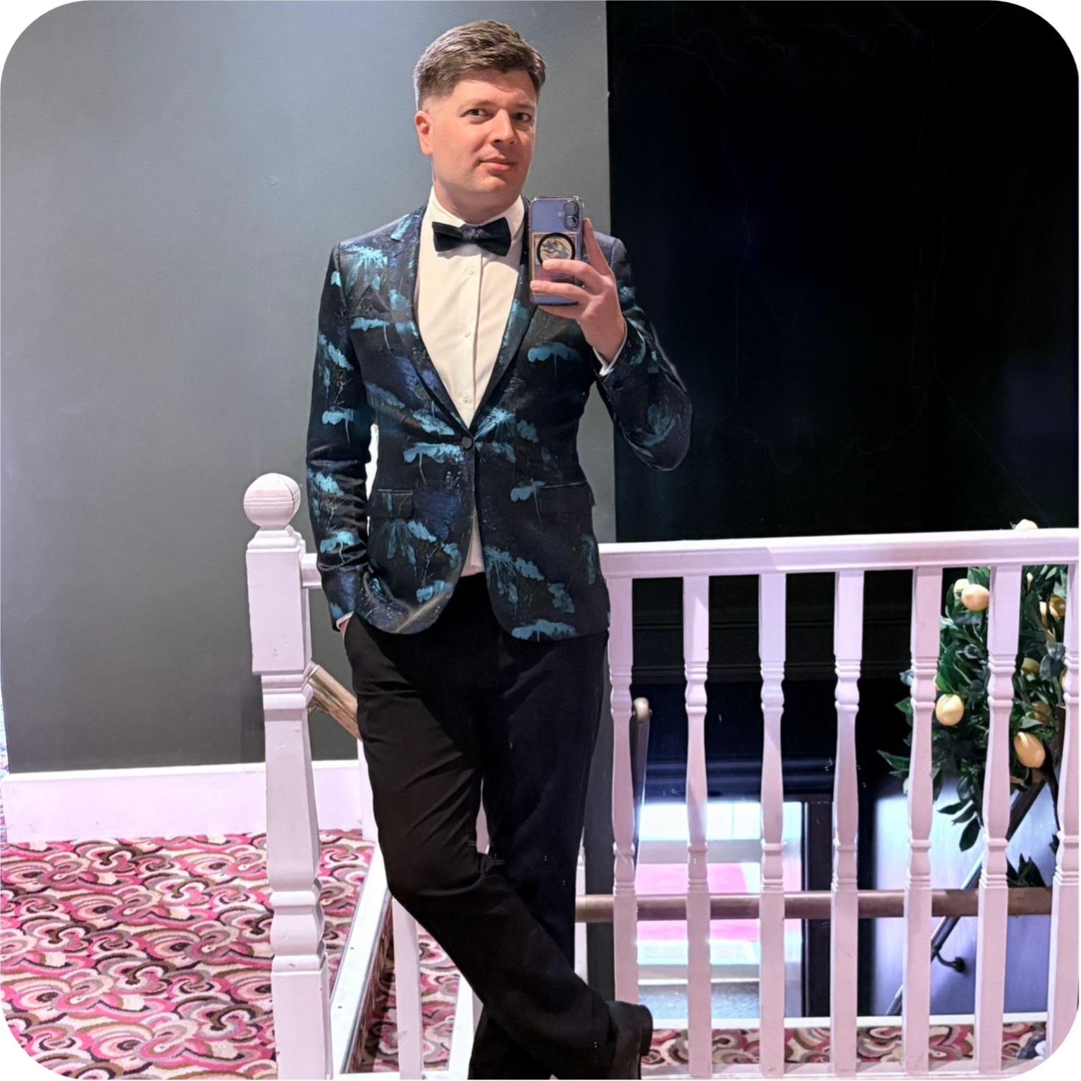 Matthew, a Queers of the North network member, stands at the top of a staircase taking a mirror selfie. They are wearing a patterned tuxedo.