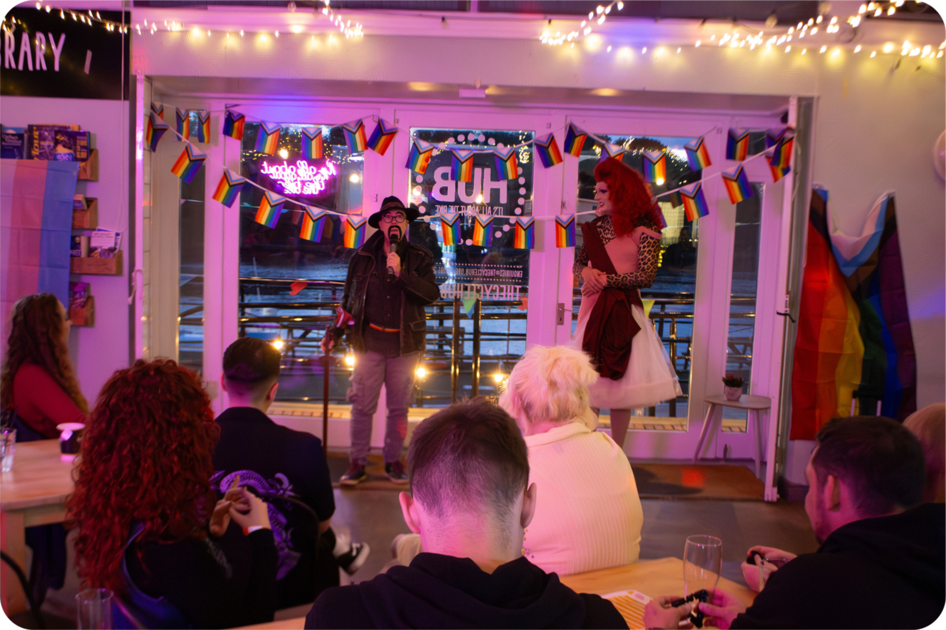North East drag artists, Sir Phoenix and Phoebe LaReine, stand front and centre at The Queer Coffee Club: After Hours event. They are surrounded by a crowd in a dimly lit setting, with LGBTQIA+ bunting and flags, and fairy lights.