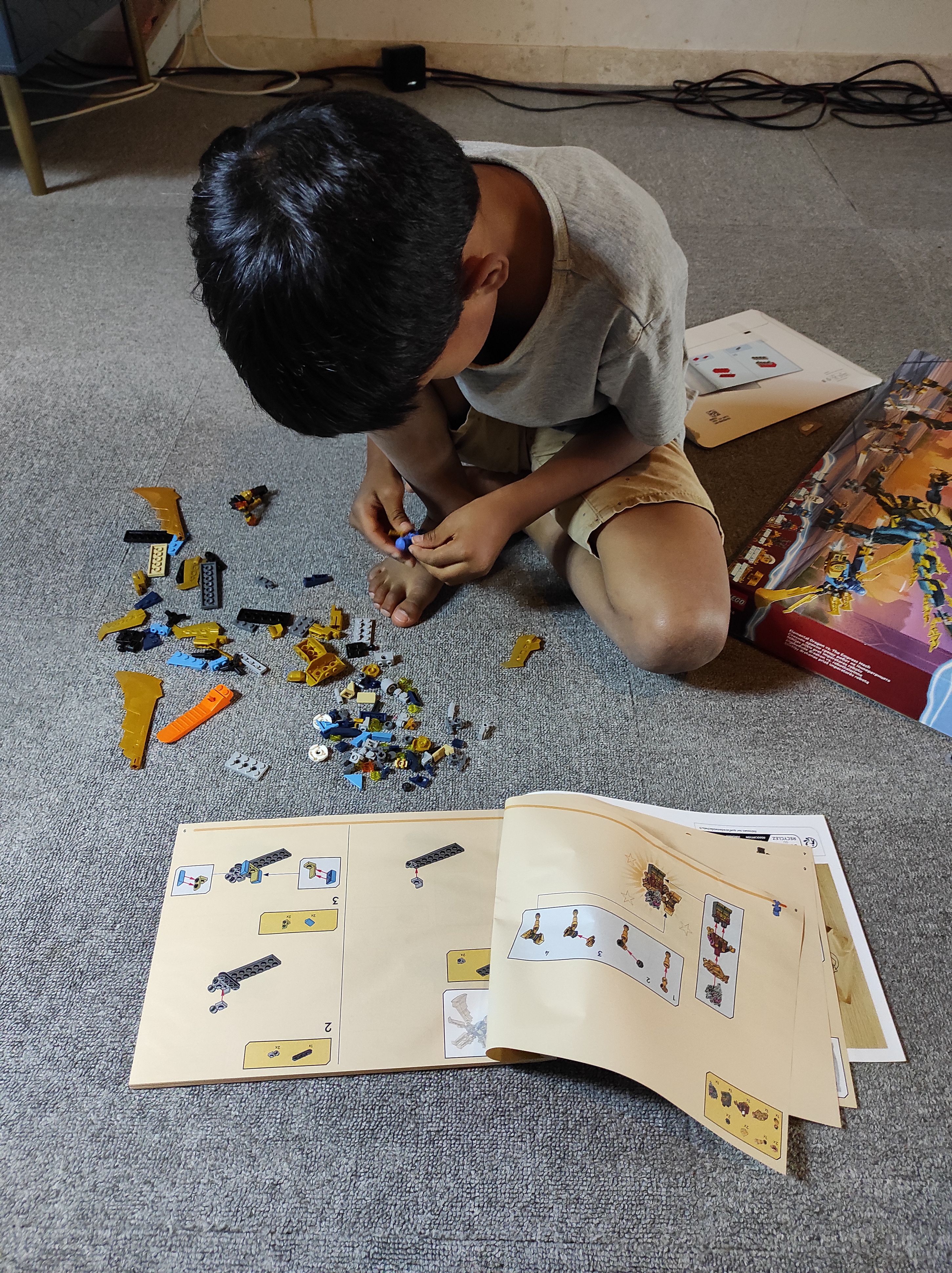 RUN is focused on building the Ninjago Dragons Rising, Elemental Dragon vs. The Empress Mech LEGOⓇ set. He’s sitting on the floor and is bending forward while clicking 2 LEGOⓇ bricks together. You can see the top of his head covered with straight, black hair and he’s wearing a grey shirt and light brown shorts. In front of him are scattered LEGOⓇ bricks and in front of is the open instruction manual. The box and packaging is visible on the right of the image.
