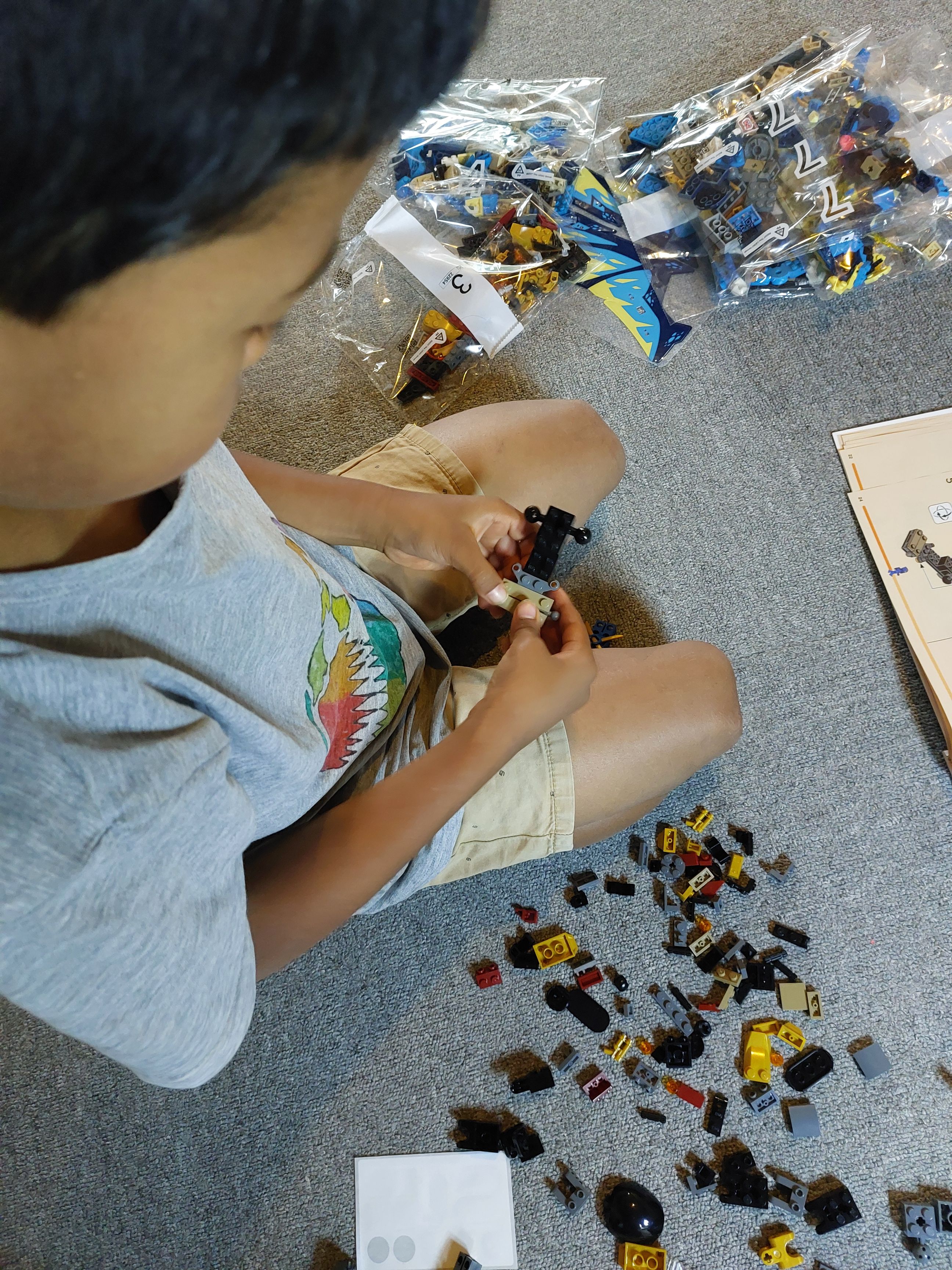 RUN is focused on building the Ninjago Dragons Rising, Elemental Dragon vs. The Empress Mech LEGOⓇ set. He’s crouching on the floor wearing a light grey t-shirt with a colorful image in the front and light brown shorts. This time, the angle is looking down at him from the right side. To his left are closed packets of LEGO<sup>Ⓡ</sup> bricks, to his right are scattered LEGOⓇ bricks. In front of him you can just see the left corner of the instruction manual.