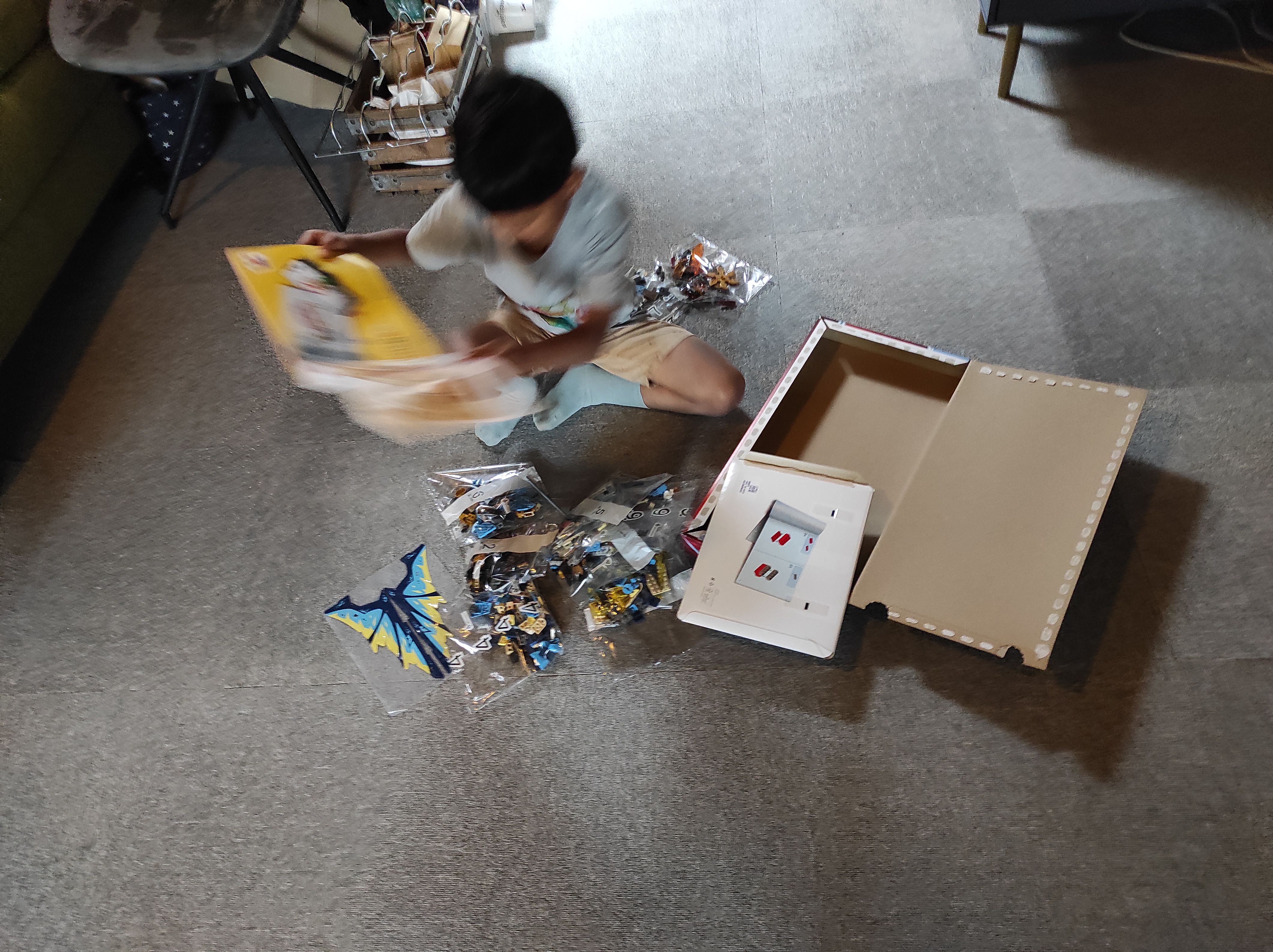 RUN excitedly unpacks the Ninjago Dragons Rising, Elemental Dragon vs. The Empress Mech LEGOⓇ set. He’s sitting on the ground wearing a light-colored t-shirt, shorts and socks and holding the instruction manual. The top half of his body and the instruction manual are blurred because of the movement when the picture was taken  The bags with LEGOⓇ bricks are scattered in front and behind him and the empty box sits on the right of the picture with the packaging of the instruction manual balancing on the edge closest to you.
