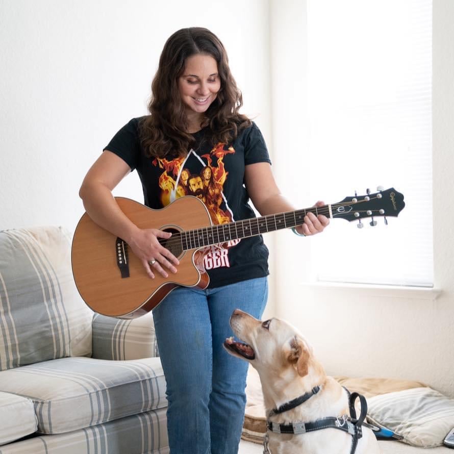 Katie playing a guitar with her retired guide dog Bristol looking up at her.