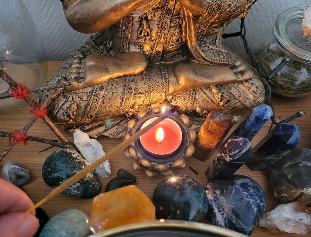 Close up photo of altar. The bottom part of a sitting buddha statue with a red tea candle lit, while an incense stick is catching fire. Around is various crystals and wooden pentagrams.