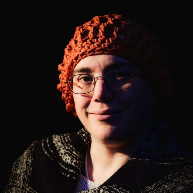 Headshot of a Caucasian woman, smiling and looking straight at the camera. Half of her face is in shadow.She is wearing a soft red-brown cap, dark blue coat, and wire-frame glasses.