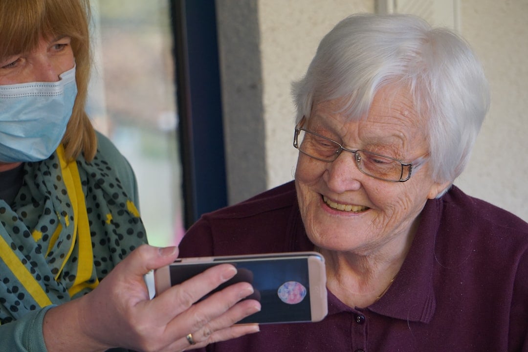 An old woman on the right, smiling while looking at the screen of a phone held by an attendant on the left.