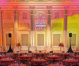 A lighting design in pink, orange, and yellow illuminates the columns and wall behind the stage in a historic ballroom.