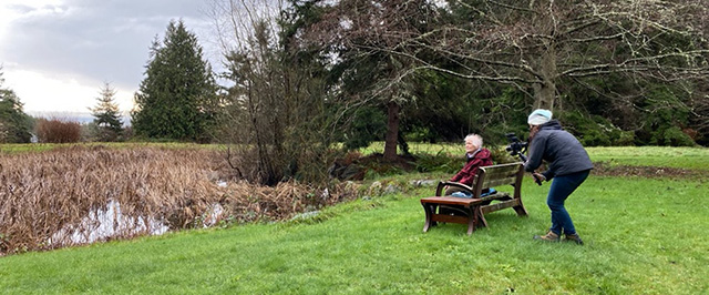 A person being filmed for a nonprofit video sits on a bench in an outdoor setting while a videographer films from behind the bench.